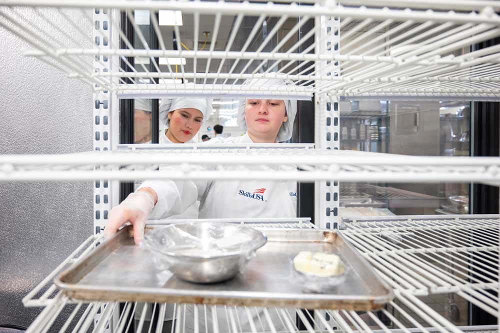 Two cooking students reaching into a rack to grab a tray